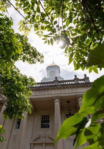 Sunlight filters through leafy green branches, framing the front of Wilbur Cross Building with its white columns and central cupola on a bright summer day.