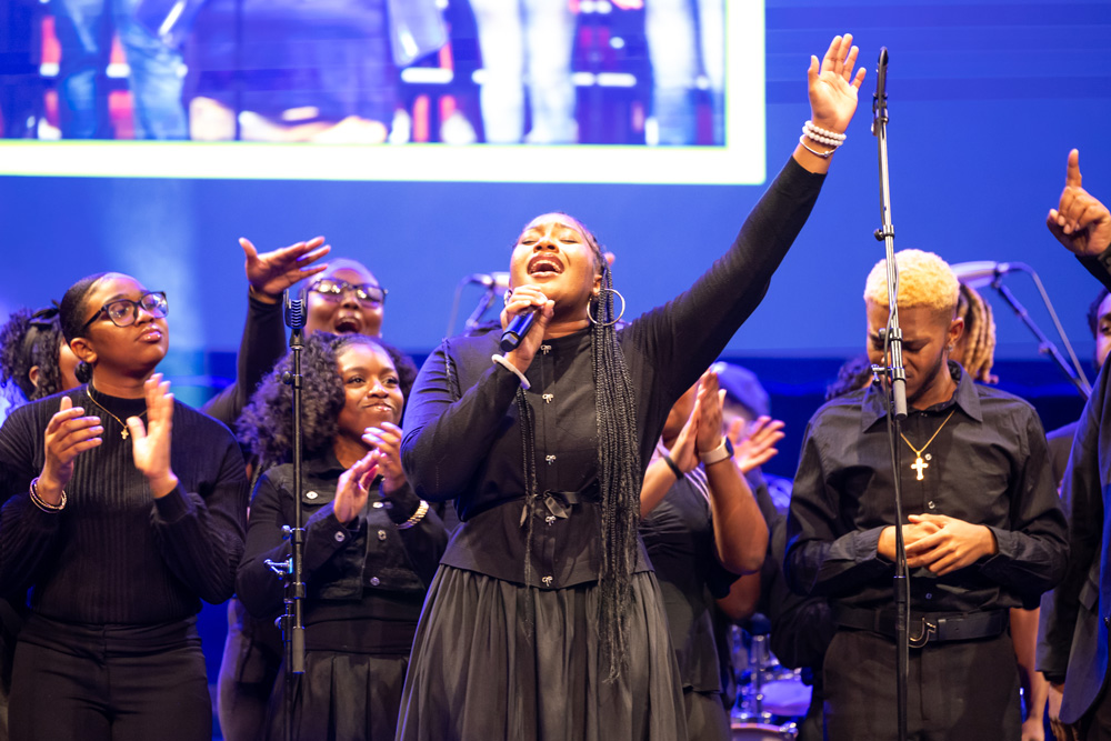 A group of performers dressed in coordinated black attire stands onstage during a musical performance, with one person at the center holding a microphone and raising an arm.