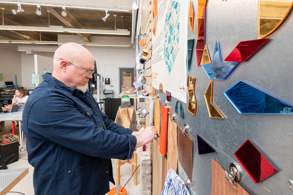 A person stands in a materials workshop examining sample swatches on a display wall filled with colorful acrylic shapes, fabric pieces, and textured materials.