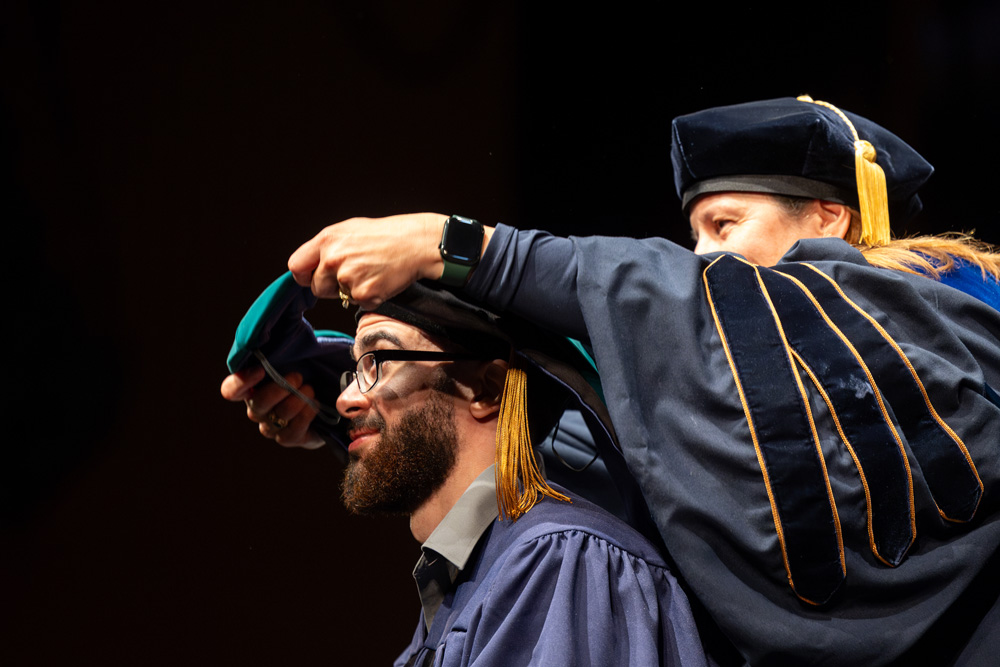 A person in academic regalia places a ceremonial hood over a graduate wearing a cap and gown during a commencement ceremony.