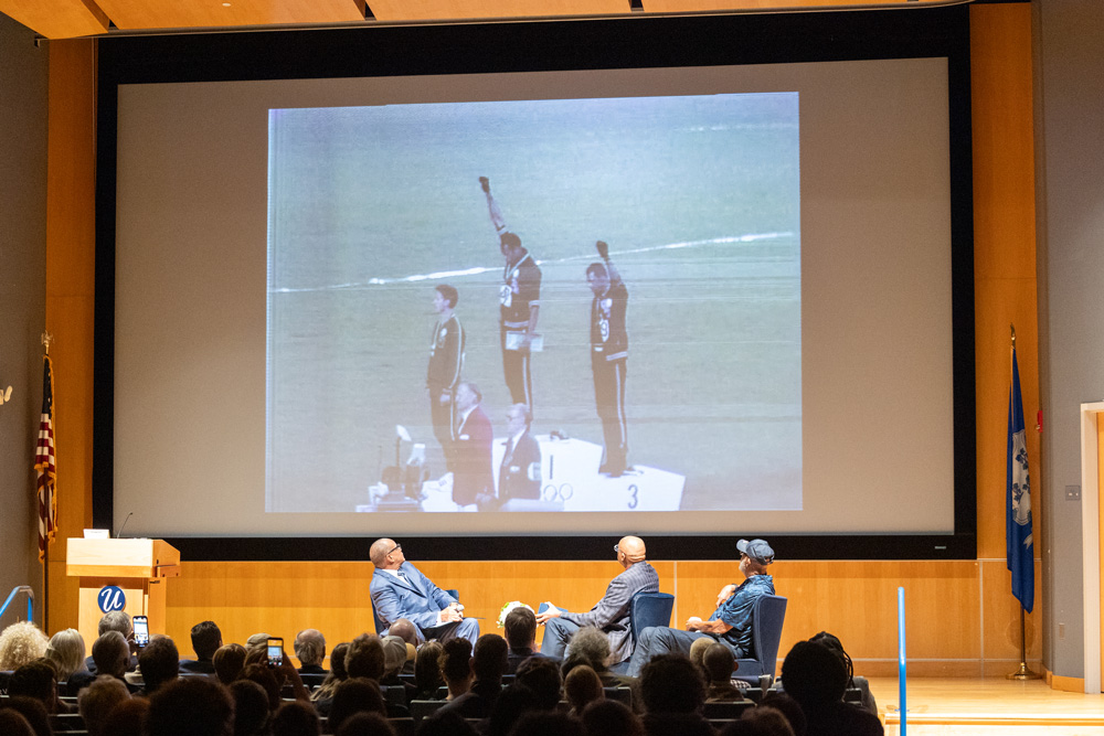 An audience watches a panel discussion in an auditorium as three speakers seated onstage view a large projected image of athletes on an Olympic medal podium.