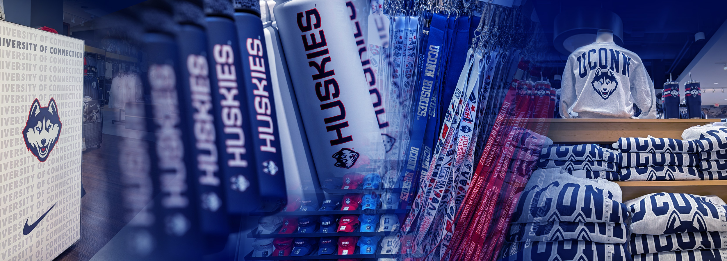 Display of UConn Huskies merchandise inside a campus store, including a large box with the Husky logo, rows of navy and white 'Huskies' scarves or apparel, racks of UConn-branded lanyards in blue, white, and red, and shelves stacked with folded UConn sweatshirts beneath a mannequin wearing a grey UConn hoodie.