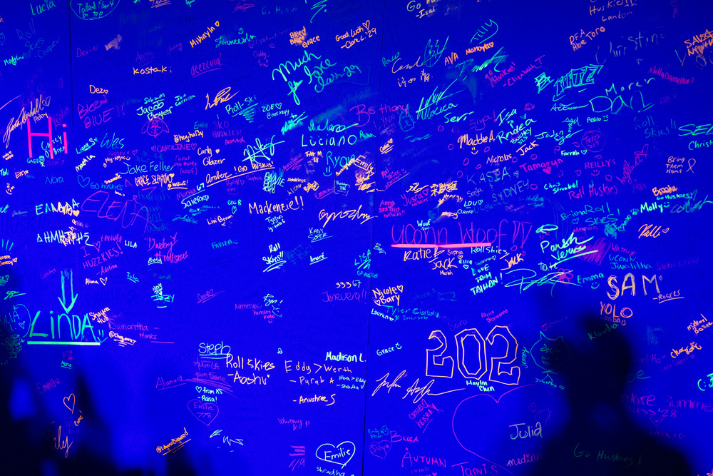 Neon-colored signatures and messages glowing under blacklight on a wall at a UConn event.
